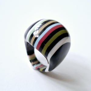 May include: A chunky dome ring featuring a smooth, polished finish with horizontal stripes in black, white, tan, light blue, red, and olive green. The ring has a wide, rounded band design that wraps around the finger with a glossy, reflective surface.