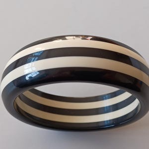 Könnte beinhalten: Ein schwarz-weiß gestreiftes Armreif-Armband. Der Armreif ist aus Kunststoff und hat eine glänzende Oberfläche.