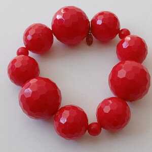 May include: A red beaded bracelet with large, faceted beads. The beads are all the same size and shape, and they are strung together on a clear elastic cord.