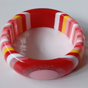 Op de afbeelding: Een rode, witte, roze en gele gestreepte armband met een roze cirkel en zilveren glitter.