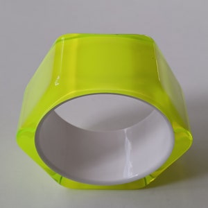 Könnte beinhalten: Ein neongrünes, geometrisches Armreif-Armband mit weißem Innenraum. Das Armband hat eine sechseckige Form und eine glänzende Oberfläche, wodurch ein modernes Accessoire entsteht. Der Durchmesser des Armbands beträgt ca. 6,35 cm.