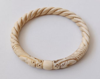 May include: A white ivory bangle bracelet with a twisted rope design and gold accents. The bracelet has carved animal heads on each side of a round bead.