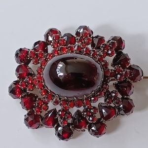 Może przedstawiać: Duża, ciemnoczerwona broszka z granatu z centralnym kaboszonem otoczonym mniejszymi granatami. Broszka jest osadzona w metalu w kolorze złotym.