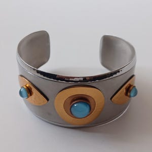 Könnte beinhalten: Silberfarbenes Armreif-Armband mit goldenen und blauen Akzenten. Das breite Armband hat eine gehämmerten Textur und ist mit goldfarbenen geometrischen Formen und hellblauen Cabochon-Steinen verziert. Ein Statement-Stück.
