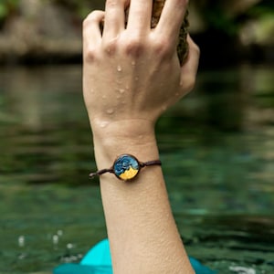 May include: A person wearing a yellow swimsuit and a blue hat with a hand raised out of the water holding a rock. The person is wearing a bracelet with a round, blue and yellow design.