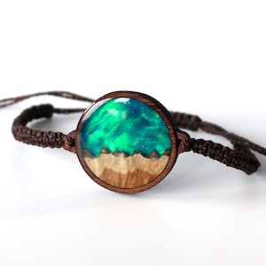May include: A brown cord bracelet with a round wooden charm featuring a green and blue resin inlay. The resin has a swirling pattern and resembles a night sky.