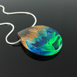 May include: A teardrop-shaped pendant necklace with a silver chain. The pendant features a wood grain top, transitioning to a vibrant blue and green resin design. The resin has a glowing effect, creating a unique and eye-catching accessory.