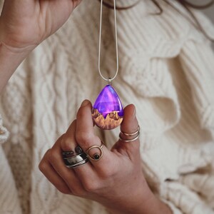 Hands holding a medium teardrop pendant made of wood, resin, and purple lab-grown opal with iridescent colors that shift from purple to pink, blue, teal, and more.