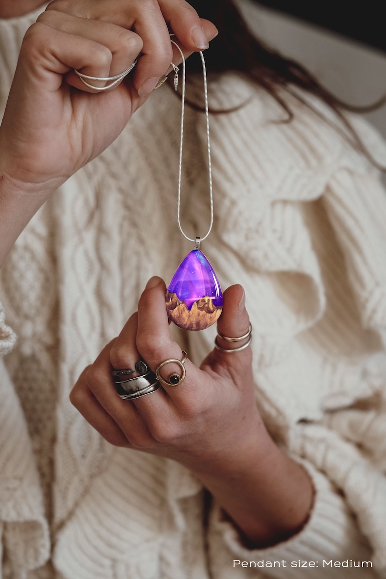 Hands holding a medium teardrop pendant made of wood, resin, and purple lab-grown opal with iridescent colors that shift from purple to pink, blue, teal, and more.