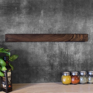 May include: A dark brown wooden magnetic knife rack mounted on a gray textured wall. The rack is empty.