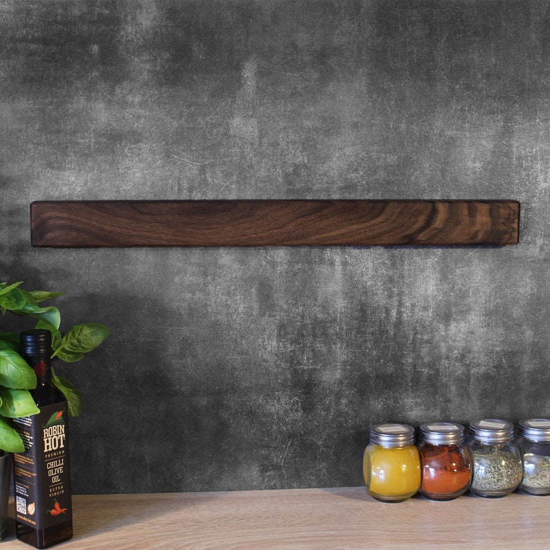 May include: A dark brown wooden magnetic knife rack mounted on a gray textured wall. The rack is empty.