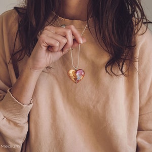 Woman wearing a heart shaped red Aurora Borealis pendant made of wood, resin, and lab-grown opal with iridescent colors that shift from green, blue, red, orange & more.
