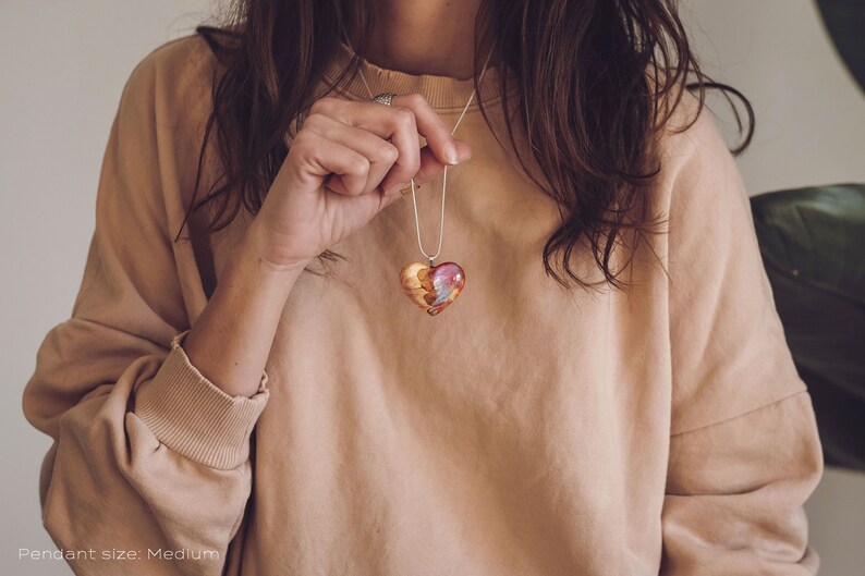 Woman wearing a heart shaped red Aurora Borealis pendant made of wood, resin, and lab-grown opal with iridescent colors that shift from green, blue, red, orange & more.