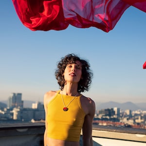 May include: A person with curly brown hair wearing a yellow tank top and a necklace with a round pendant. The person is standing on a rooftop with a city skyline in the background. A red fabric is flowing in the air above the person.