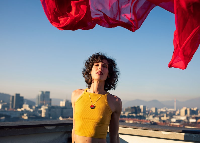 May include: A person with curly brown hair wearing a yellow tank top and a necklace with a round pendant. The person is standing on a rooftop with a city skyline in the background. A red fabric is flowing in the air above the person.
