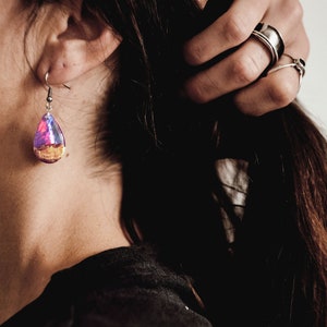 Woman wearing a teardrop purple Aurora Borealis earrings made of wood, resin, and purple lab-grown opal with iridescent colors that shift from purple to pink, blue, teal, and more.