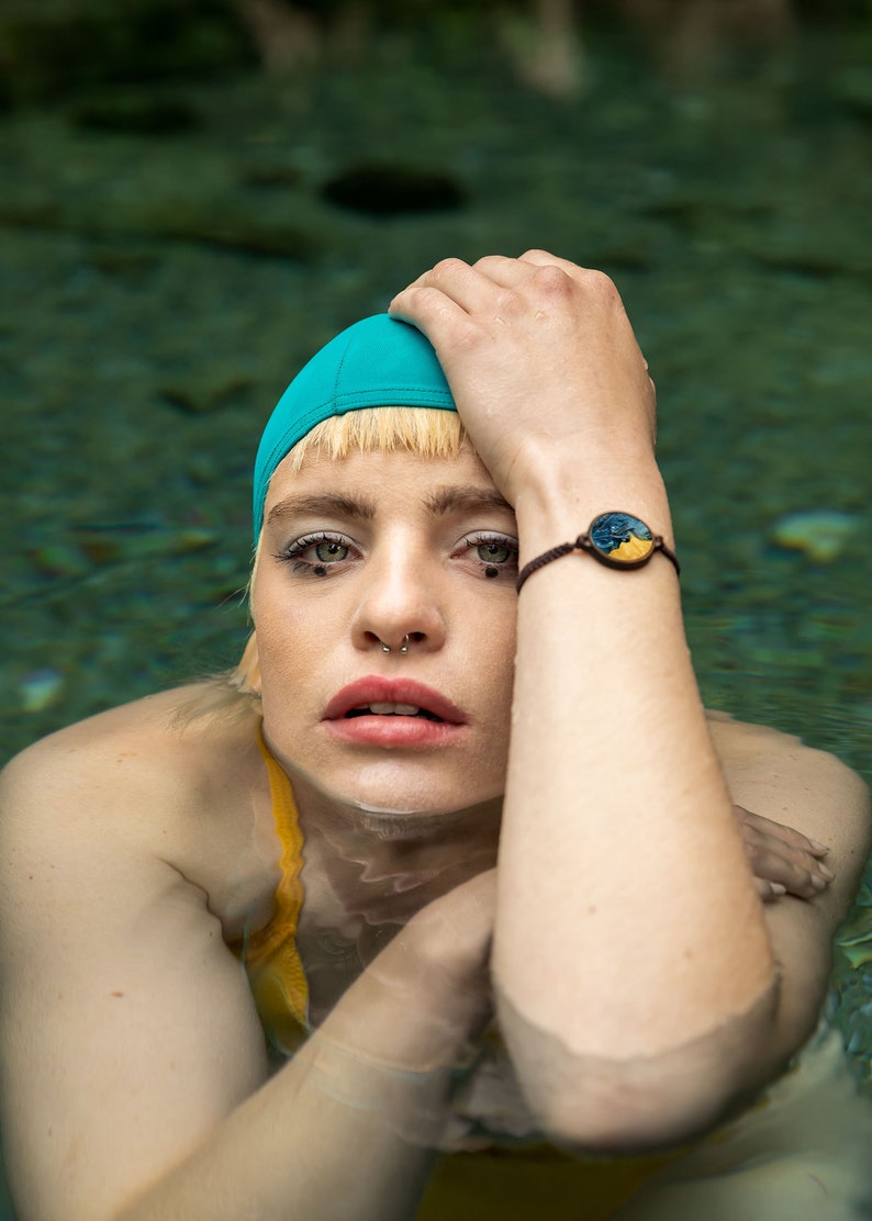 May include: A woman with blonde hair wearing a turquoise swim cap and a yellow swimsuit is submerged in a pool of water. She has a nose ring and a bracelet with a blue and yellow circular design on her wrist.