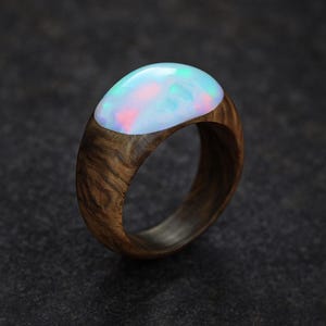 May include: A wooden ring with a large, oval opal stone. The opal has a pearlescent sheen with hints of pink, blue, and green. The ring's band is made of dark wood, showcasing a natural grain pattern. A unique piece of jewelry.