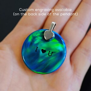 May include: A round, iridescent blue and green pendant with a silver heart charm and the words "I love you" engraved on the front.