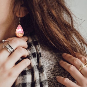 Woman wearing a teardrop pink Aurora Borealis earrings made of wood, resin, and purple lab-grown opal with iridescent colors that shift from pink to green, red and more.