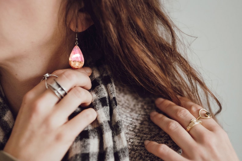 Woman wearing a teardrop pink Aurora Borealis earrings made of wood, resin, and purple lab-grown opal with iridescent colors that shift from pink to green, red and more.