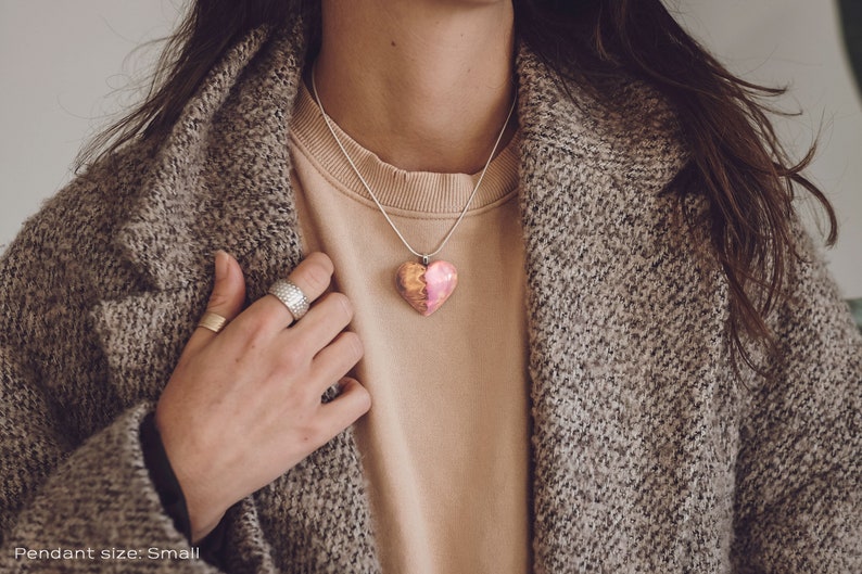 Woman wearing a heart shaped pink Aurora Borealis pendant made of wood, resin, and lab-grown opal with iridescent colors that shift from purple to pink to green, red and more.