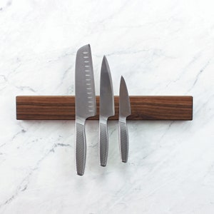 May include: A wooden magnetic knife holder with three silver knives attached to it. The holder is brown and has a natural wood grain. The knives are different sizes and have a brushed finish.