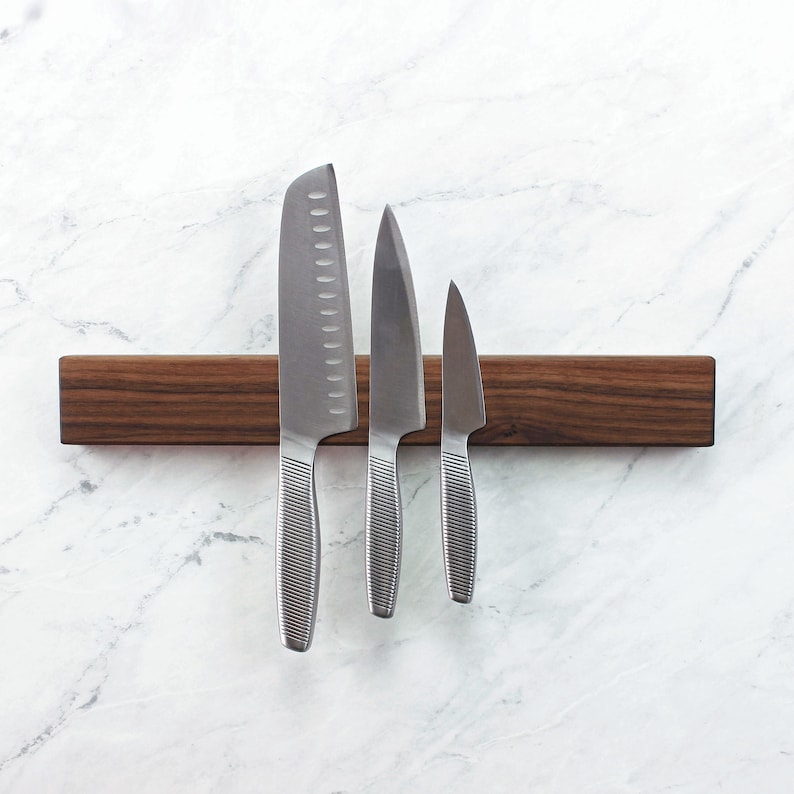 May include: A wooden magnetic knife holder with three silver knives attached to it. The holder is brown and has a natural wood grain. The knives are different sizes and have a brushed finish.