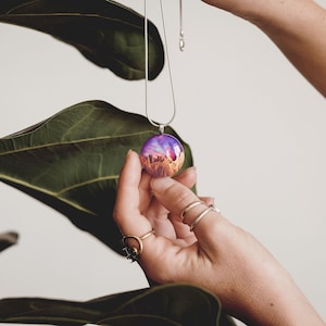 Hands holding a small round pendant made of wood, resin, and purple lab-grown opal with iridescent colors that shift from purple to pink, blue, teal, and more.