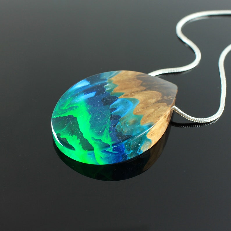 May include: A teardrop-shaped pendant necklace with a silver chain. The pendant features a wood base with a translucent resin top. The resin is layered with shades of green, blue, and brown, creating a unique, artistic design. The necklace is a piece of wearable art.