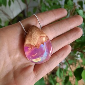 Hand holding teardrop pendant made of wood and resin, featuring pink lab-grown opal with iridescent colors changing from pink to green, red and more.