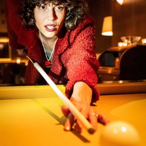 May include: A woman in a red sweater leans over a pool table, lining up a shot with a pool cue. A white billiard ball is in the foreground.
