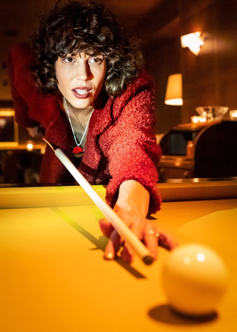 May include: A woman in a red sweater leans over a pool table, lining up a shot with a pool cue. A white billiard ball is in the foreground.