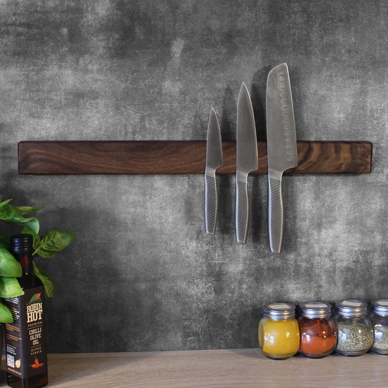 May include: A dark brown wooden magnetic knife holder with three silver knives attached to it. The holder is mounted on a gray wall. There are four small glass jars with lids on a light brown surface below the holder.
