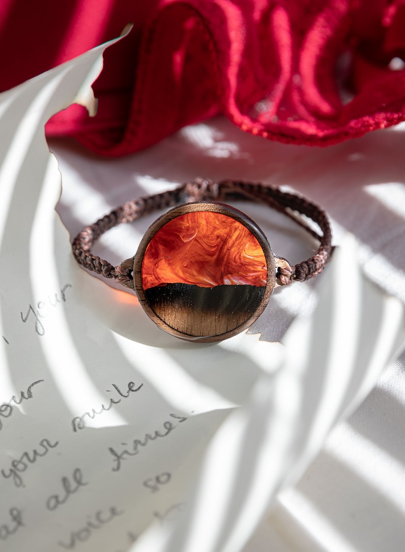 May include: A brown leather bracelet with a round wooden charm. The charm has a resin inlay with a swirling orange and black design.