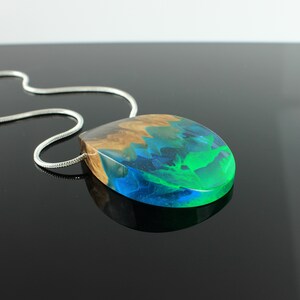 May include: A handcrafted pendant necklace featuring a teardrop-shaped resin pendant. The pendant combines wood and vibrant colors: brown, blue, and green. The pendant is suspended from a silver chain, creating a unique and artistic accessory.