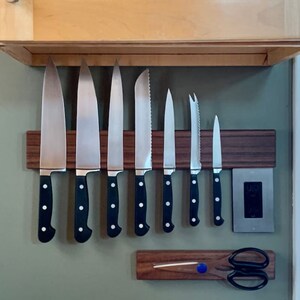 May include: A set of eight kitchen knives with black handles are magnetically attached to a wooden wall-mounted knife rack. A pair of kitchen shears are attached to a smaller wooden rack below the knife rack.