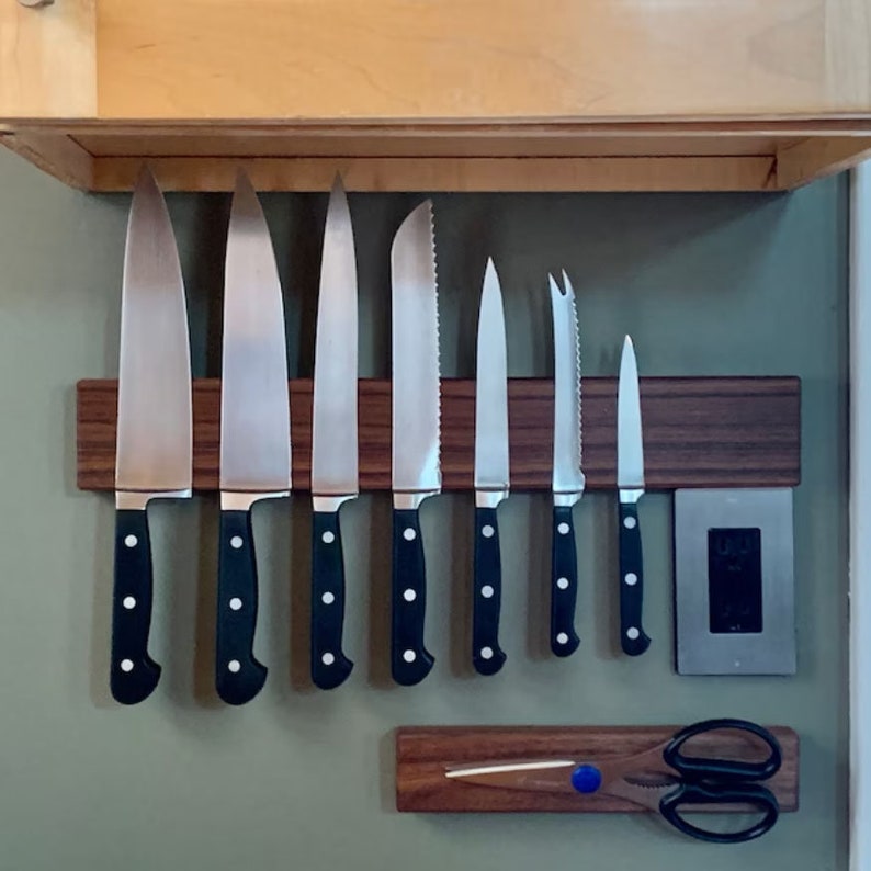 May include: A set of eight kitchen knives with black handles are magnetically attached to a wooden wall-mounted knife rack. A pair of kitchen shears are attached to a smaller wooden rack below the knife rack.