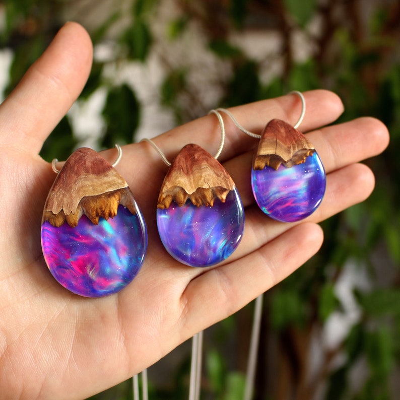 Hand holding three wood and resin teardrop pendants of varying sizes - small, medium and large, each featuring purple lab-grown opal with iridescent colors changing from purple to pink, blue, teal, and more.
