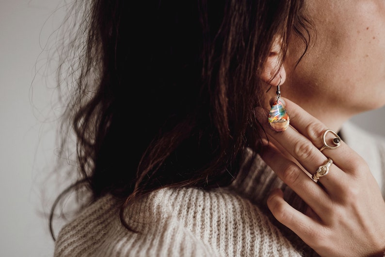 Woman wearing a teardrop red Aurora Borealis earrings made of wood, resin, and purple lab-grown opal with iridescent colors that shift from green, blue, red, orange & more.