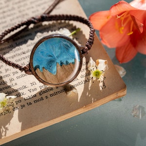 May include: A brown leather bracelet with a round wooden charm. The charm has a blue and white resin design that resembles a seascape.