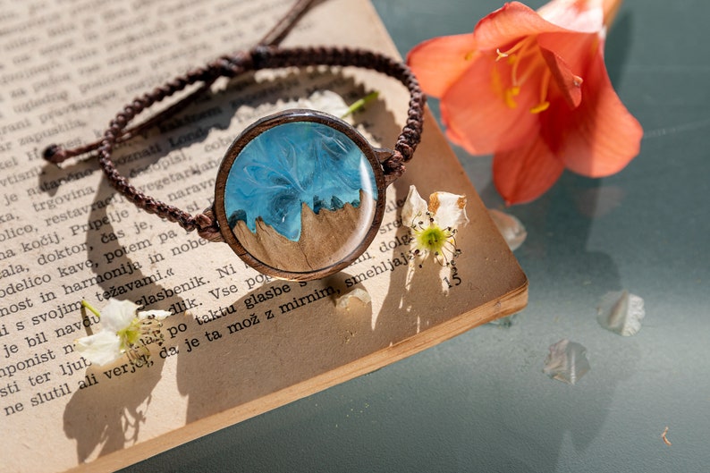 May include: A brown leather bracelet with a round wooden charm. The charm has a blue and white resin design that resembles a seascape.
