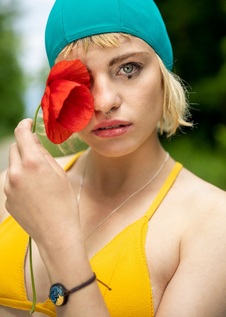 May include: A woman with blonde hair is wearing a turquoise swim cap and a yellow bikini top. She is holding a red poppy flower in front of her eye. She has a nose ring and a bracelet on her wrist.