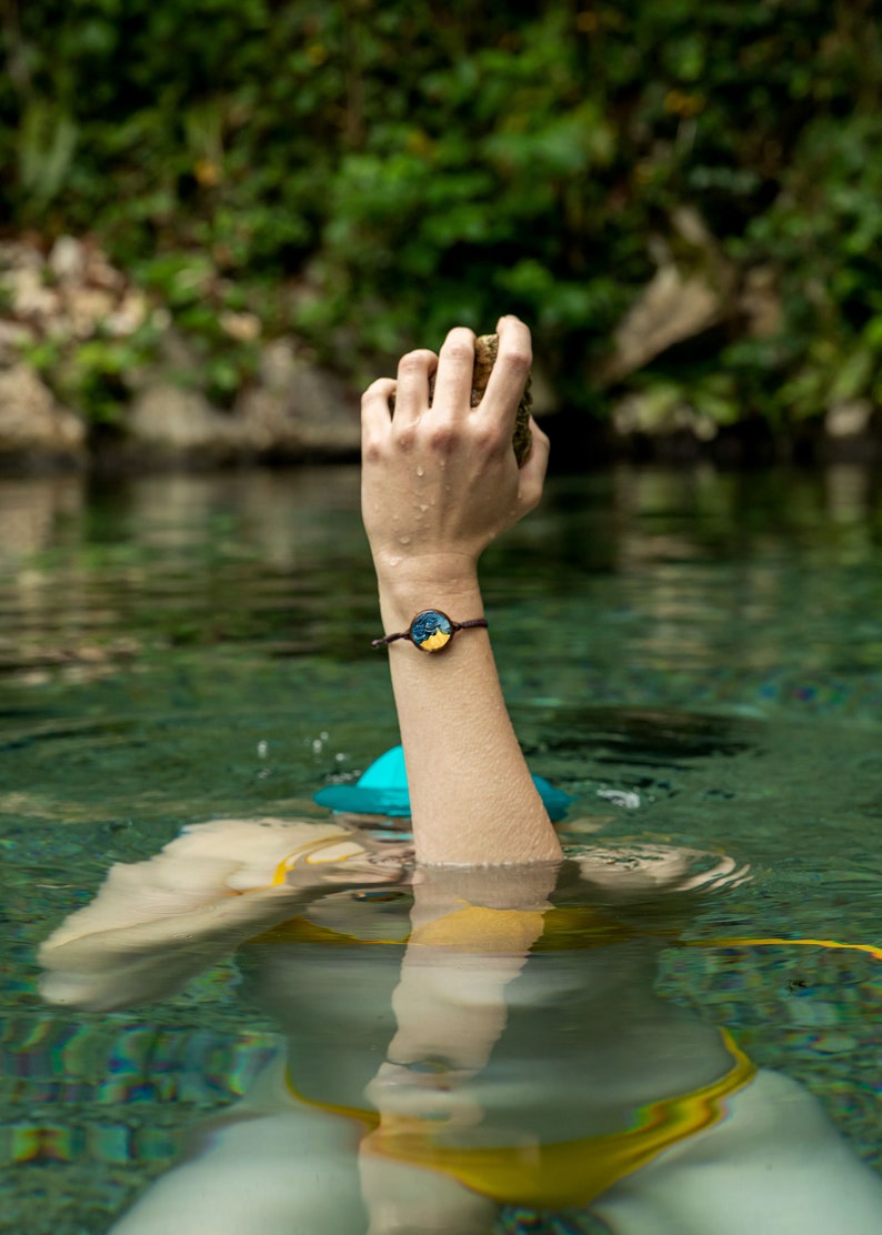 May include: A person wearing a yellow swimsuit and a blue hat with a hand raised out of the water holding a rock. The person is wearing a bracelet with a round, blue and yellow design.