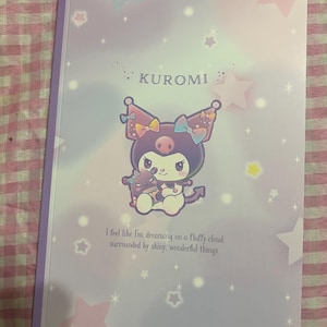May include: A notebook with a light purple cover featuring the character Kuromi. The cover has a starry design and the text "KUROMI". The image includes the text: "I feel like I'm dreaming on a fluffy cloud, surrounded by shiny, wonderful things."
