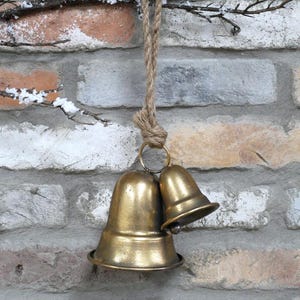 May include: Two antique brass bells suspended from a natural rope against a brick wall. The bells, of different sizes, have a warm, golden hue. The brick wall features a mix of colours, including white, grey, and brown.