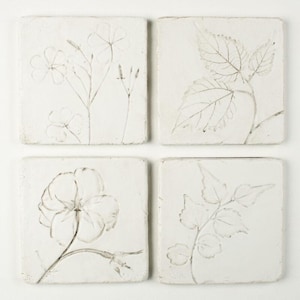 May include: Four square, off-white coasters with embossed floral designs. Each coaster features a different botanical illustration in a muted grey tone. The designs include flowers and leaves, creating a natural, artistic aesthetic.