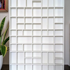 May include: A white wall-mounted display unit with a grid of cubby holes. The shelf is made of wood and painted white. The design is modern and suitable for displaying small items or organizing objects.