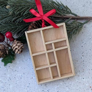 May include: A small, wooden, rectangular ornament with multiple compartments. It has a red ribbon for hanging. The ornament is displayed with pine needles, pine cones, and red berries, suggesting a holiday theme.