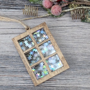 May include: Wooden Christmas ornament shaped like a window, with six panes filled with iridescent snowflake confetti. The ornament has a natural wood finish and a jute string for hanging. Festive holiday decor.